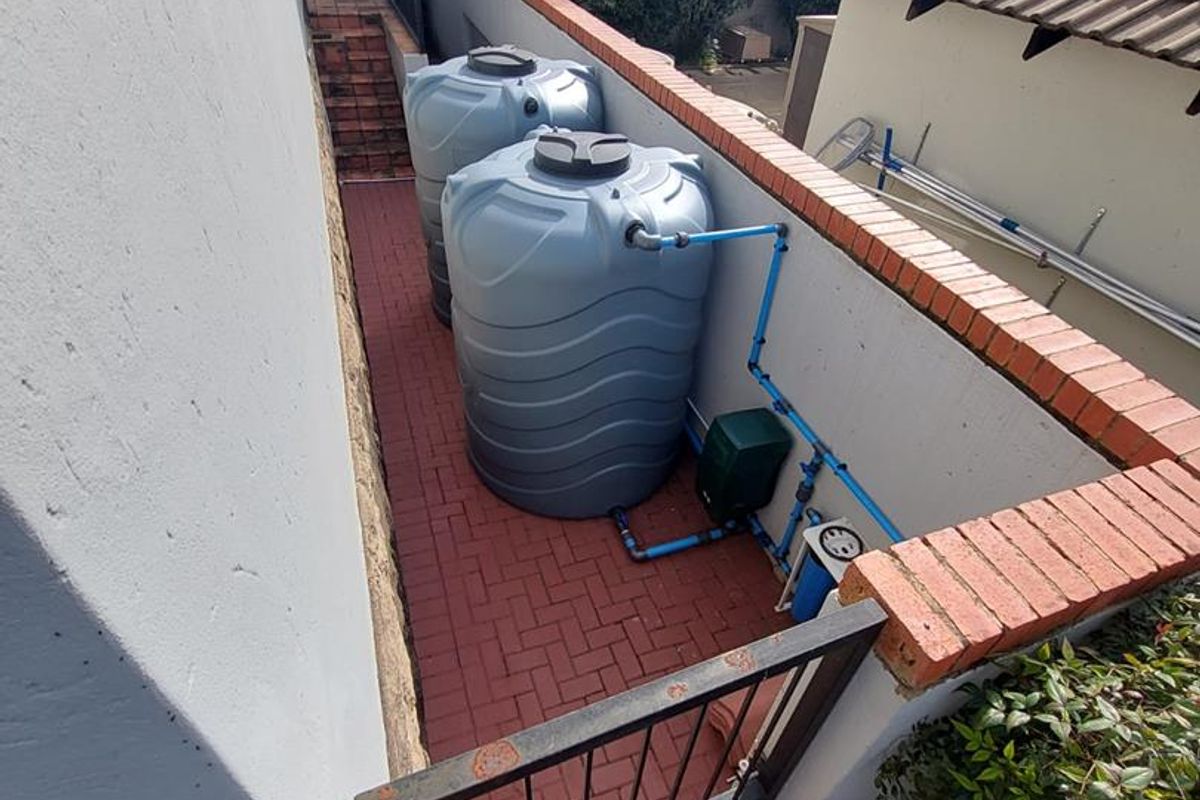 Side exterior area featuring two large water storage tanks, a pressure pump system, and paved flooring enclosed by a brick wall and metal gate.