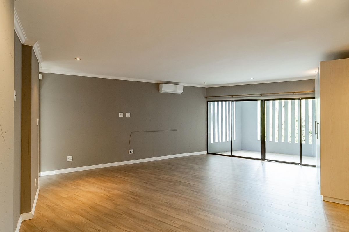 A spacious, empty living area featuring modern wood-look flooring, recessed lighting, a wall-mounted air conditioning unit, and large sliding glass doors leading to a balcony.