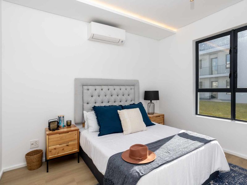 A modern, well-lit bedroom featuring laminate flooring, a wall-mounted air conditioning unit, and large windows with black aluminium frames.