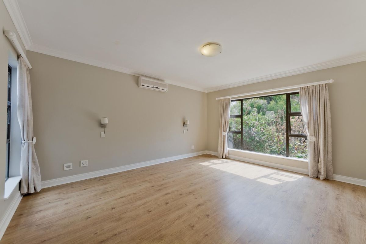 A bright and spacious bedroom featuring light wood-look laminate flooring, an air conditioning unit, and large windows with garden views.