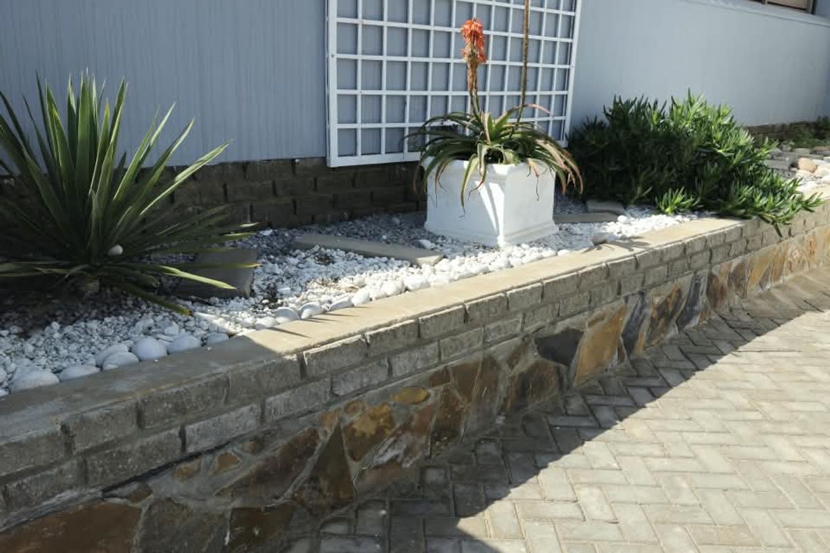 A well-maintained rear exterior area featuring a stone-clad retaining wall, paved ground surface, and a decorative trellis against the boundary wall.