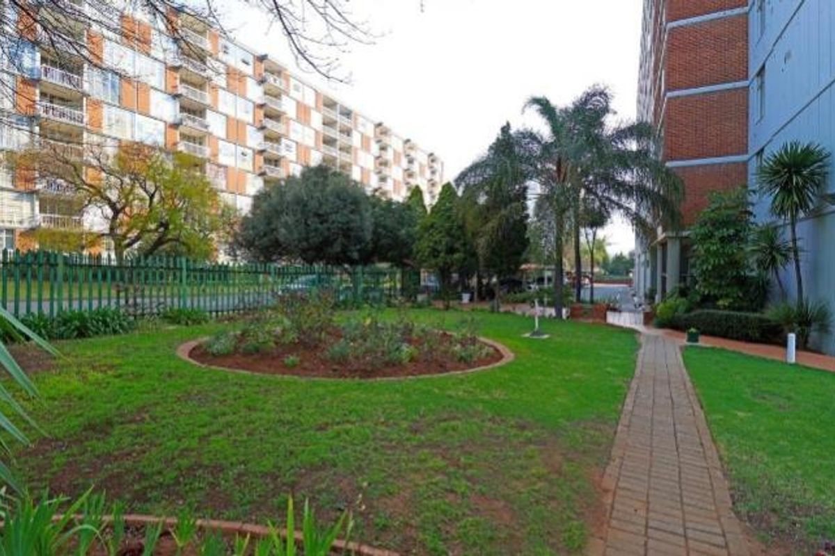 A well-maintained communal garden area featuring a paved walkway, established trees, and landscaped beds situated between apartment blocks.