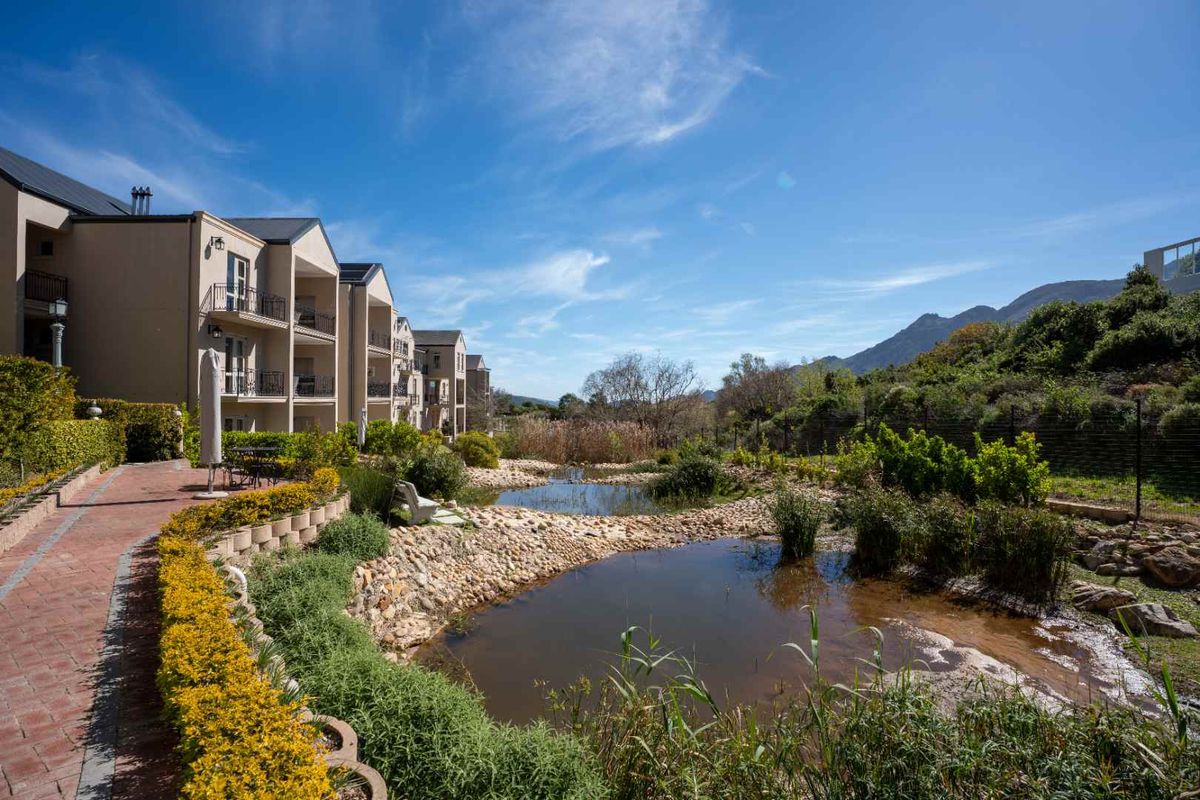 A scenic view of the apartment complex exterior featuring a landscaped garden, stone-lined ponds, and a brick paved walkway leading to the building.