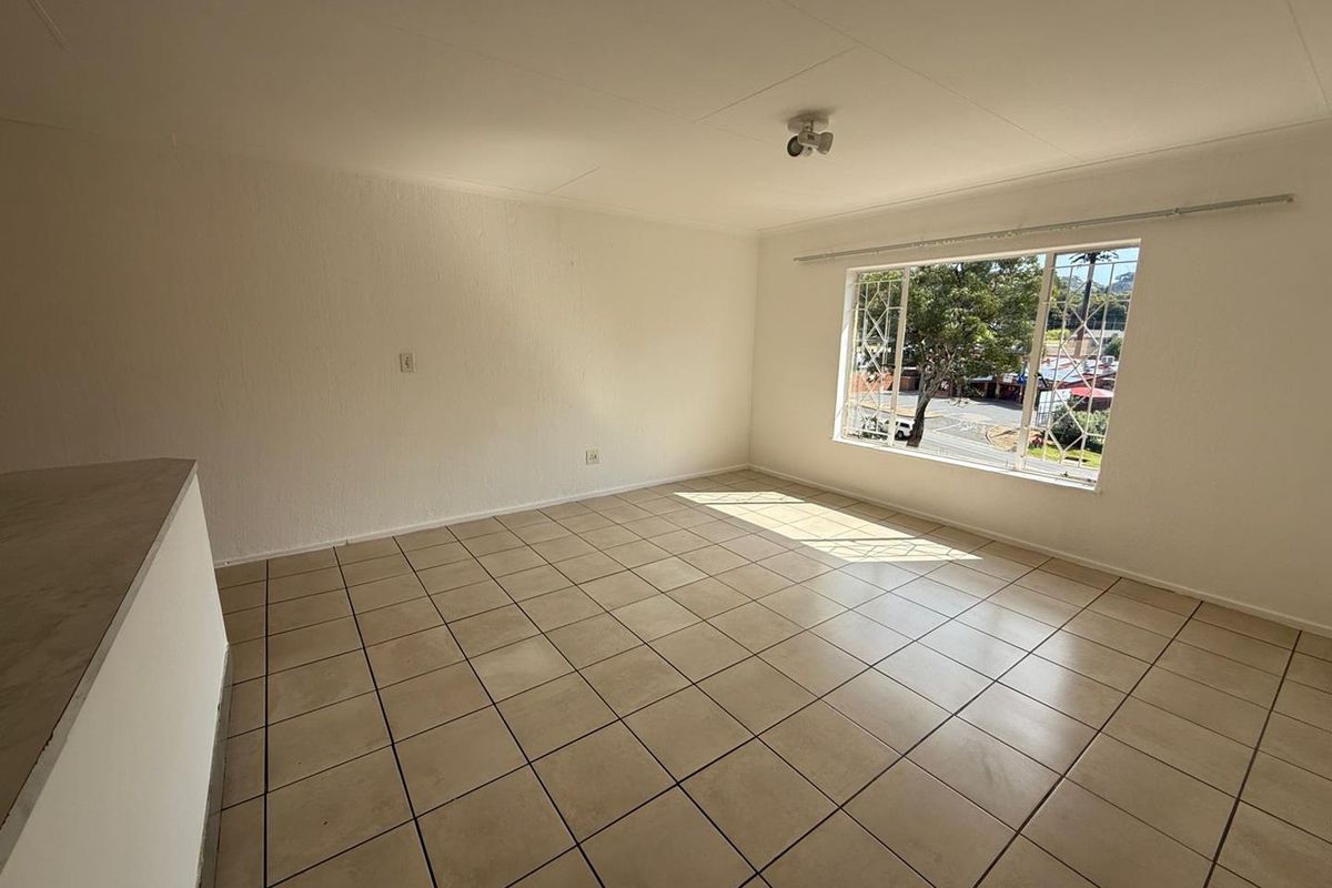 An empty, sunlit living area featuring neutral tiled flooring and a large window with security bars, offering a blank canvas for interior design.