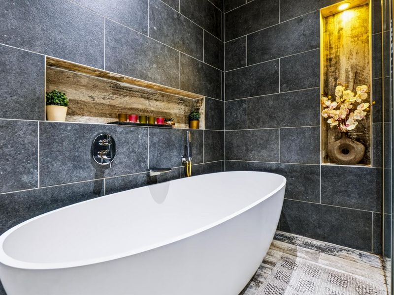 Modern bathroom featuring a sleek freestanding bathtub, dark stone wall tiling, and elegant recessed niches with integrated accent lighting.
