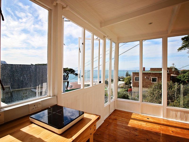 A bright, enclosed sunroom or balcony area featuring timber flooring, extensive glass windows, and panoramic coastal views.