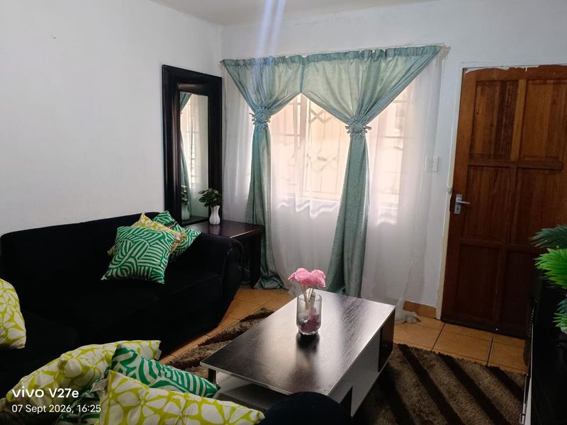 A compact living room featuring tiled flooring, a wooden entrance door, and a window fitted with security bars.