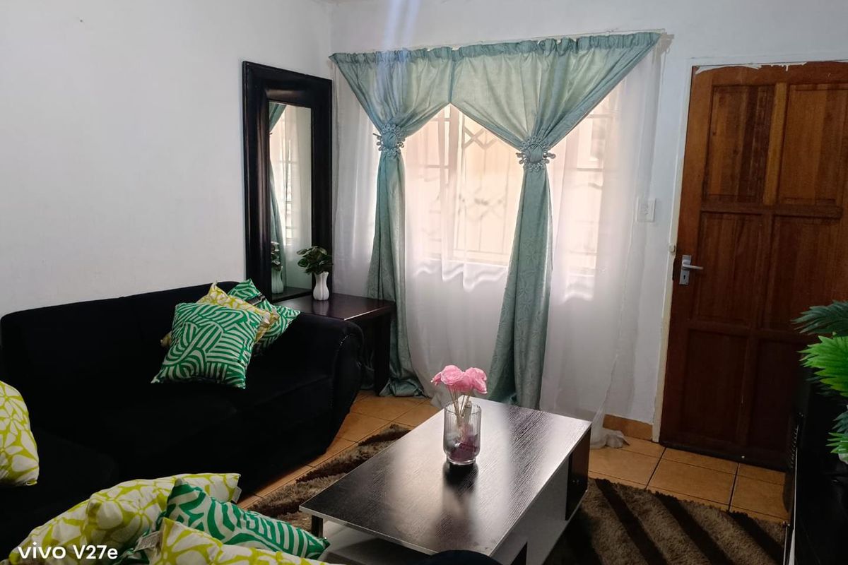 A compact living room featuring tiled flooring, a wooden entrance door, and a window fitted with security bars.