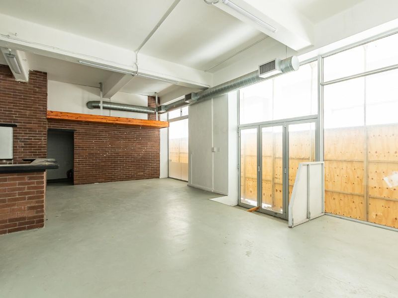Spacious open-plan industrial style area featuring exposed brickwork, high ceilings with exposed ducting, and large windows currently boarded with plywood.