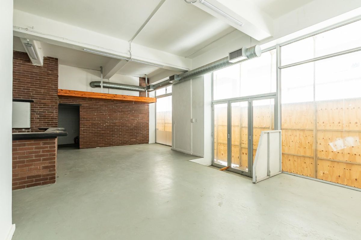 Spacious open-plan industrial style area featuring exposed brickwork, high ceilings with exposed ducting, and large windows currently boarded with plywood.