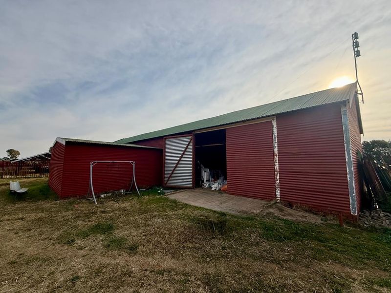 A large red corrugated metal farm utility building featuring a sliding barn door and concrete apron, set on a rural property.
