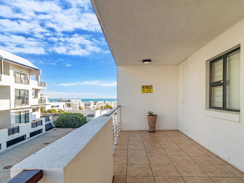 A spacious covered balcony featuring tiled flooring, a metal balustrade, and views of the surrounding apartment complex and distant ocean.