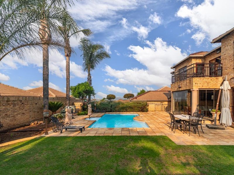 A well-maintained backyard featuring a swimming pool, paved patio area, lush lawn, and a two-story brick house with a balcony.