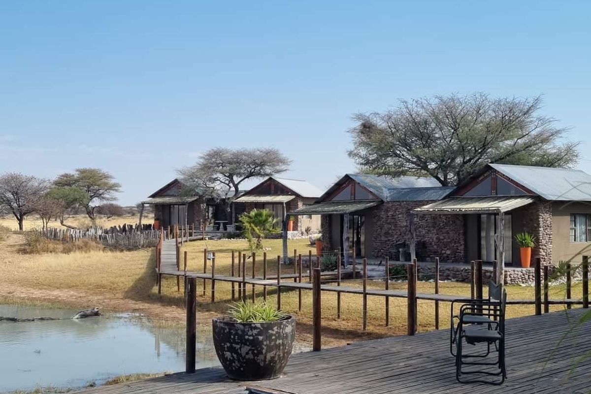 A scenic view of rustic game farm chalets featuring stone masonry, corrugated metal roofing, and elevated wooden walkways overlooking a natural waterhole.