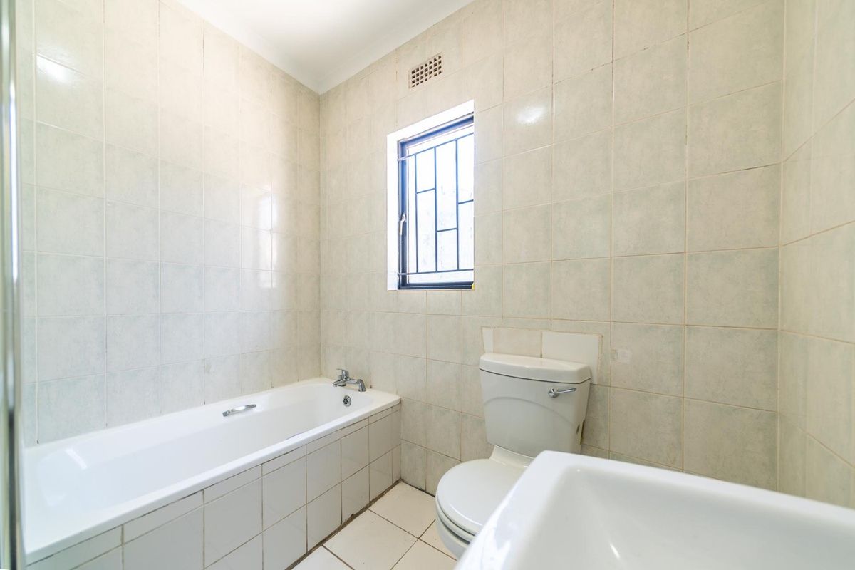 A functional bathroom featuring a bathtub, toilet, and washbasin with full-height wall tiling and a window with security bars.