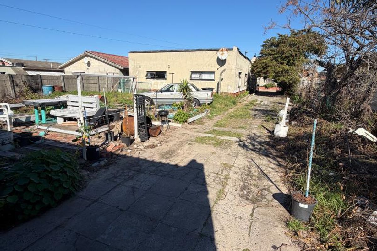 Rear exterior view of the property featuring a concrete driveway, boundary wall, and significant overgrown garden area requiring maintenance.