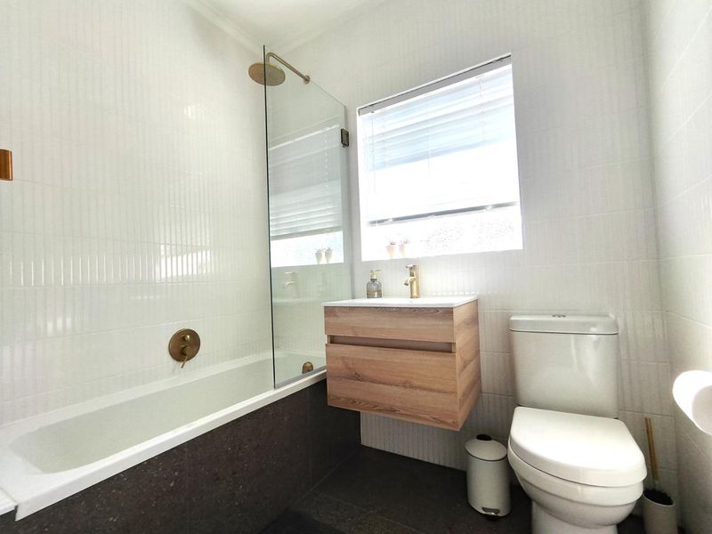Modern bathroom featuring a wall-mounted vanity, built-in bathtub with glass screen, brass fixtures, and neutral tiled finishes.