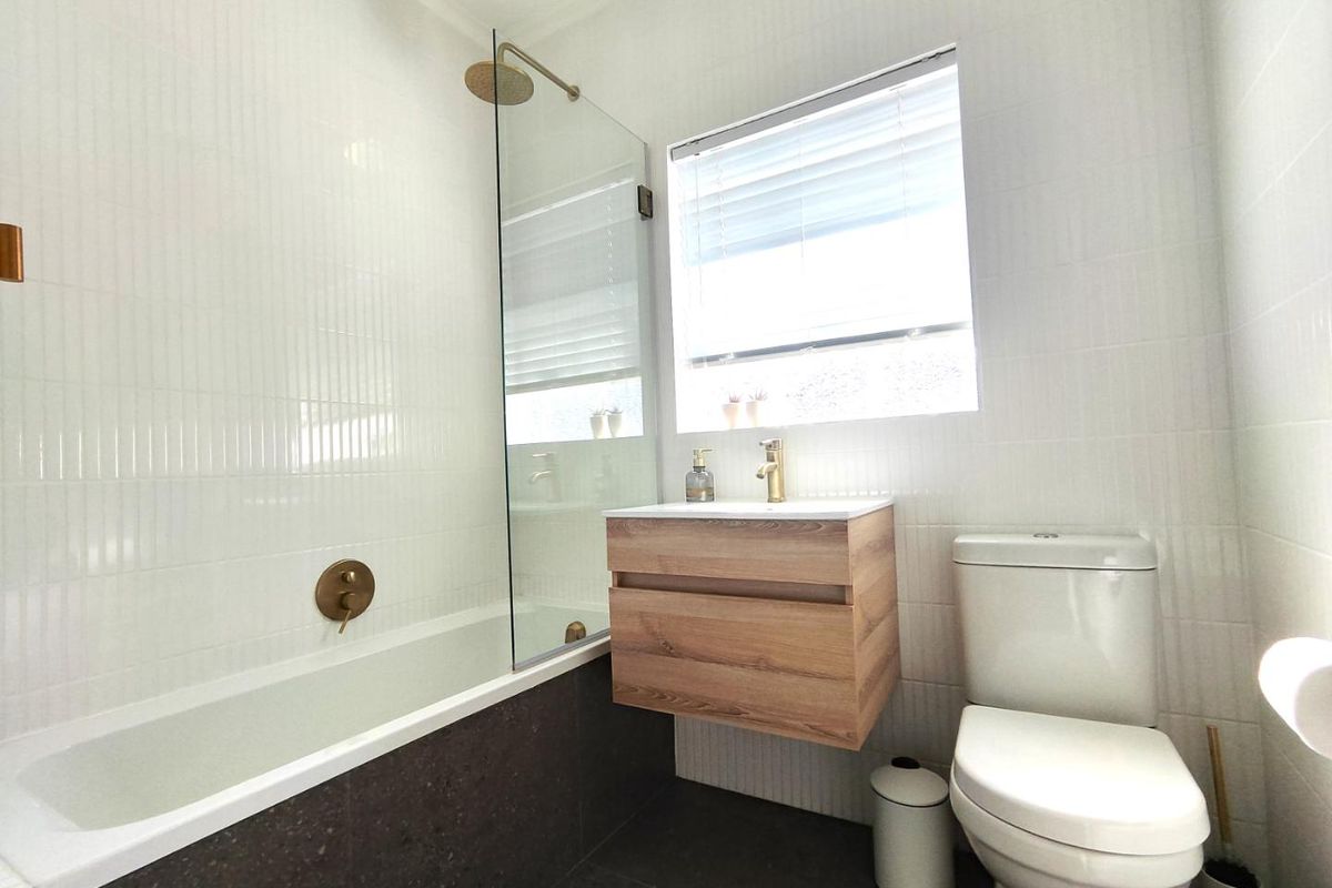 Modern bathroom featuring a wall-mounted vanity, built-in bathtub with glass screen, brass fixtures, and neutral tiled finishes.