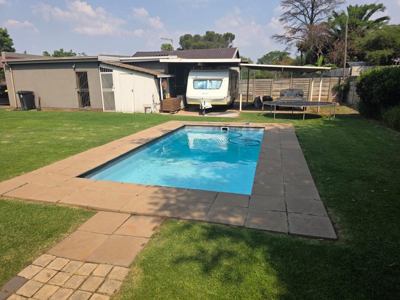 A backyard area featuring a rectangular swimming pool with paved surround, adjacent lawn, and a covered carport structure.