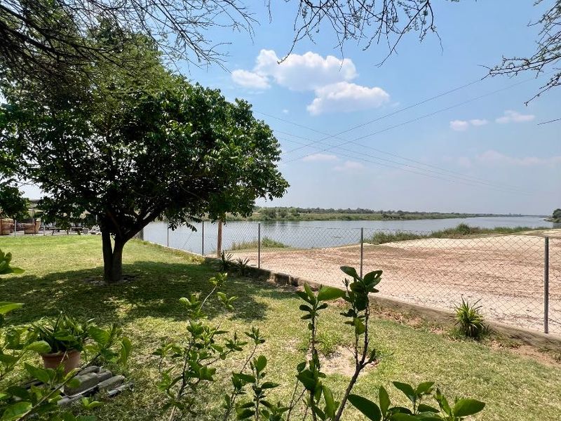 A scenic view of the riverfront area featuring a well-maintained lawn, mature trees, and secure wire mesh fencing along the property boundary.