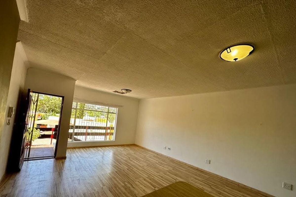 A spacious, empty living room featuring laminate flooring, textured ceilings, and natural light from a large window with security bars.