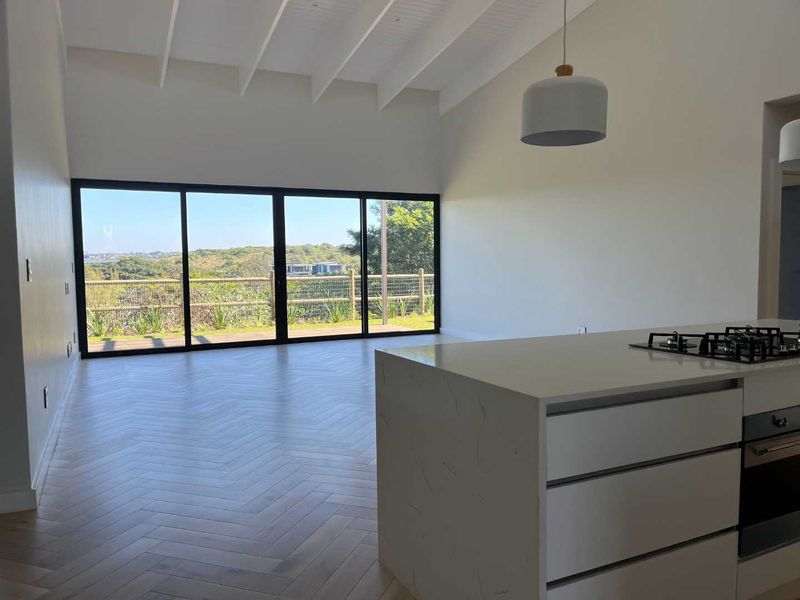Spacious open-plan living area featuring herringbone timber flooring, a vaulted ceiling, and a modern kitchen island with built-in appliances, opening to scenic views.