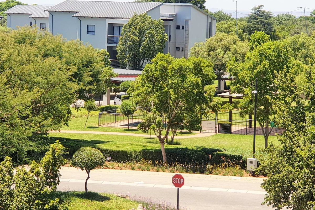 An elevated aerial view of the apartment complex grounds, featuring landscaped gardens, a fenced pool area, and surrounding mature trees.
