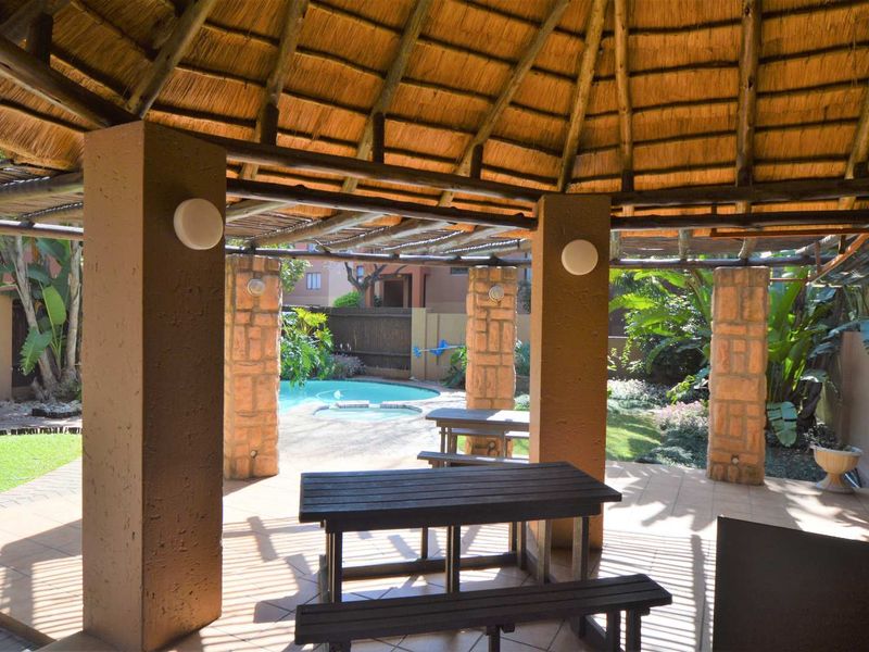 A covered outdoor patio area featuring a traditional thatched roof, stone-clad support pillars, and tiled flooring, overlooking a private garden and swimming pool.