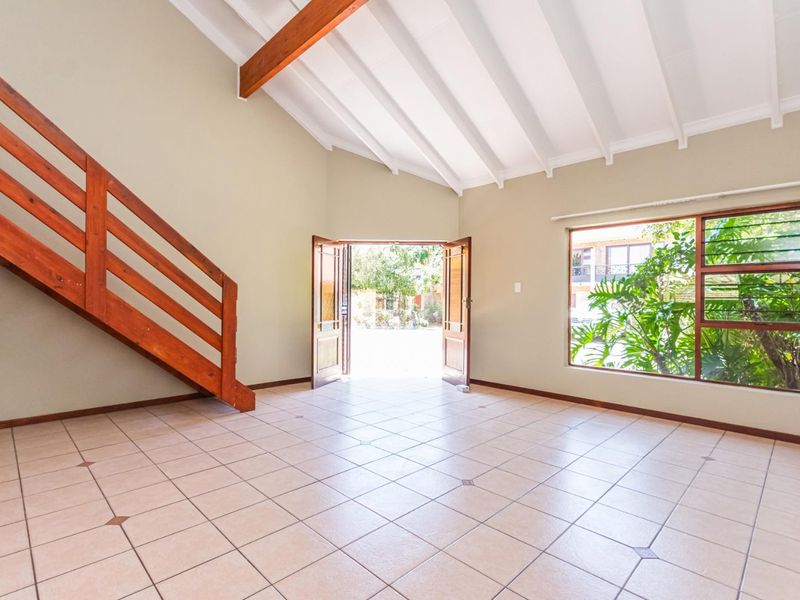 Spacious living area featuring high vaulted ceilings with exposed timber beams, tiled flooring, and a wooden staircase leading to an upper level.