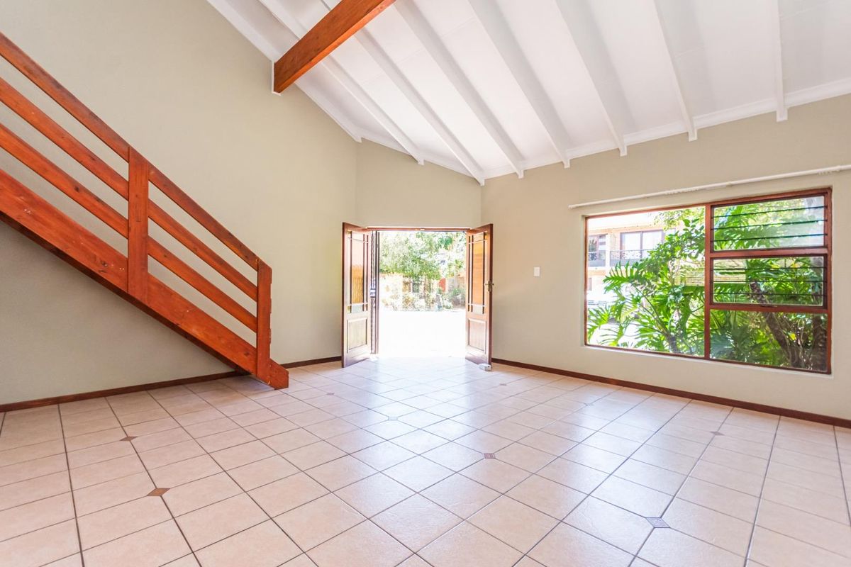 Spacious living area featuring high vaulted ceilings with exposed timber beams, tiled flooring, and a wooden staircase leading to an upper level.