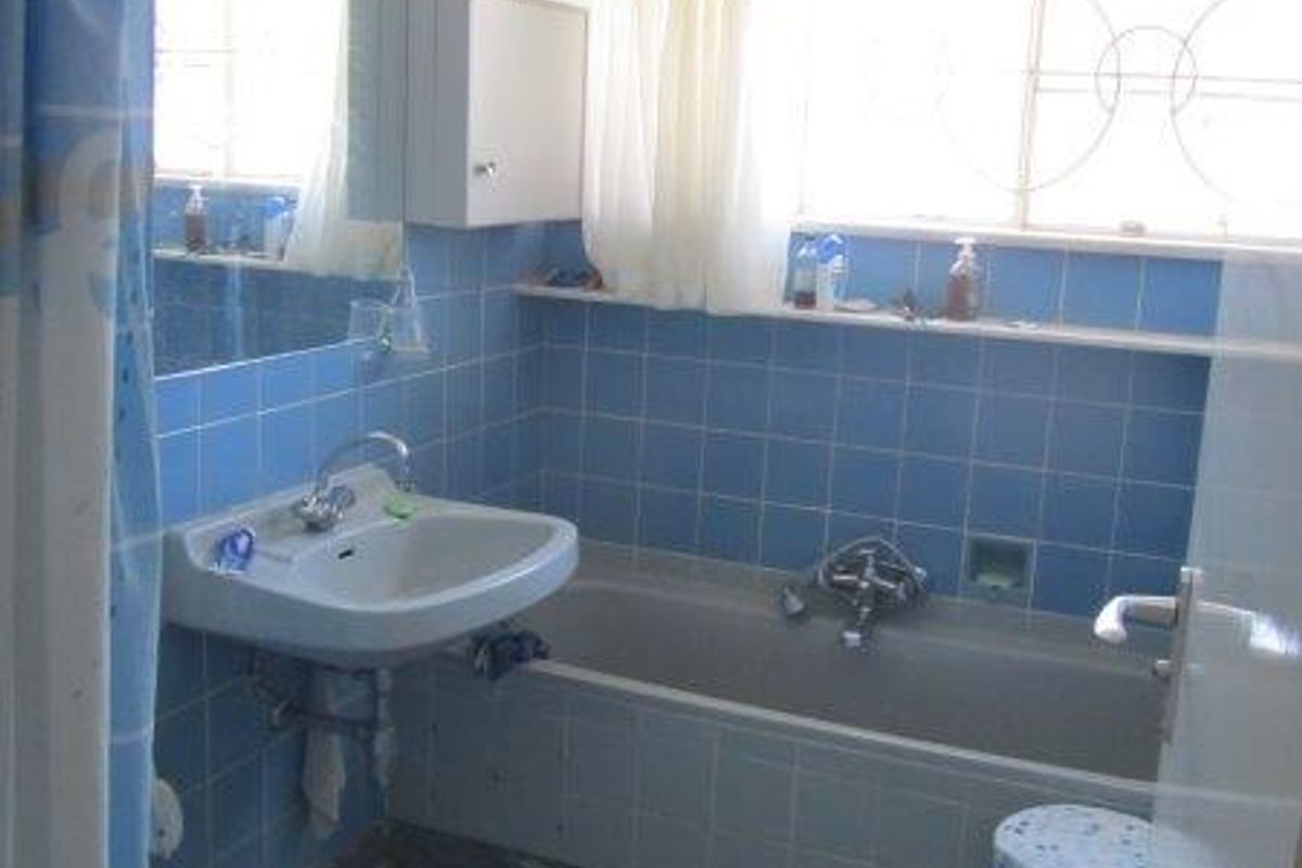 A dated bathroom featuring blue wall tiles, a built-in bathtub, a wall-mounted basin, and a window with security bars.