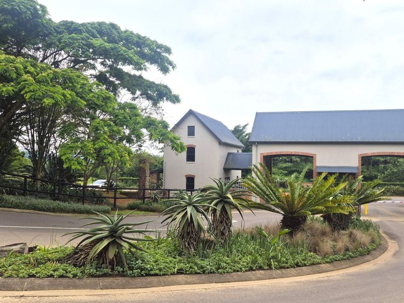 A well-maintained residential estate entrance featuring brick and plaster architecture, a metal roof, and established landscaped gardens.