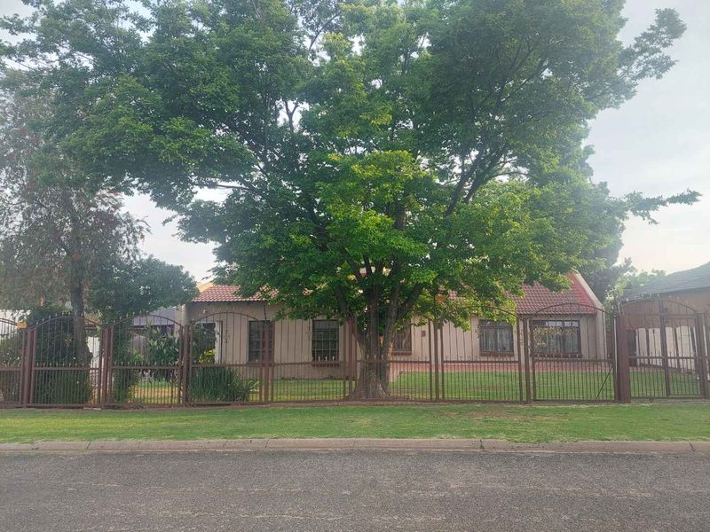 Front view of a single-story house featuring a tiled roof, secure metal perimeter fencing, and a mature tree in the front garden.