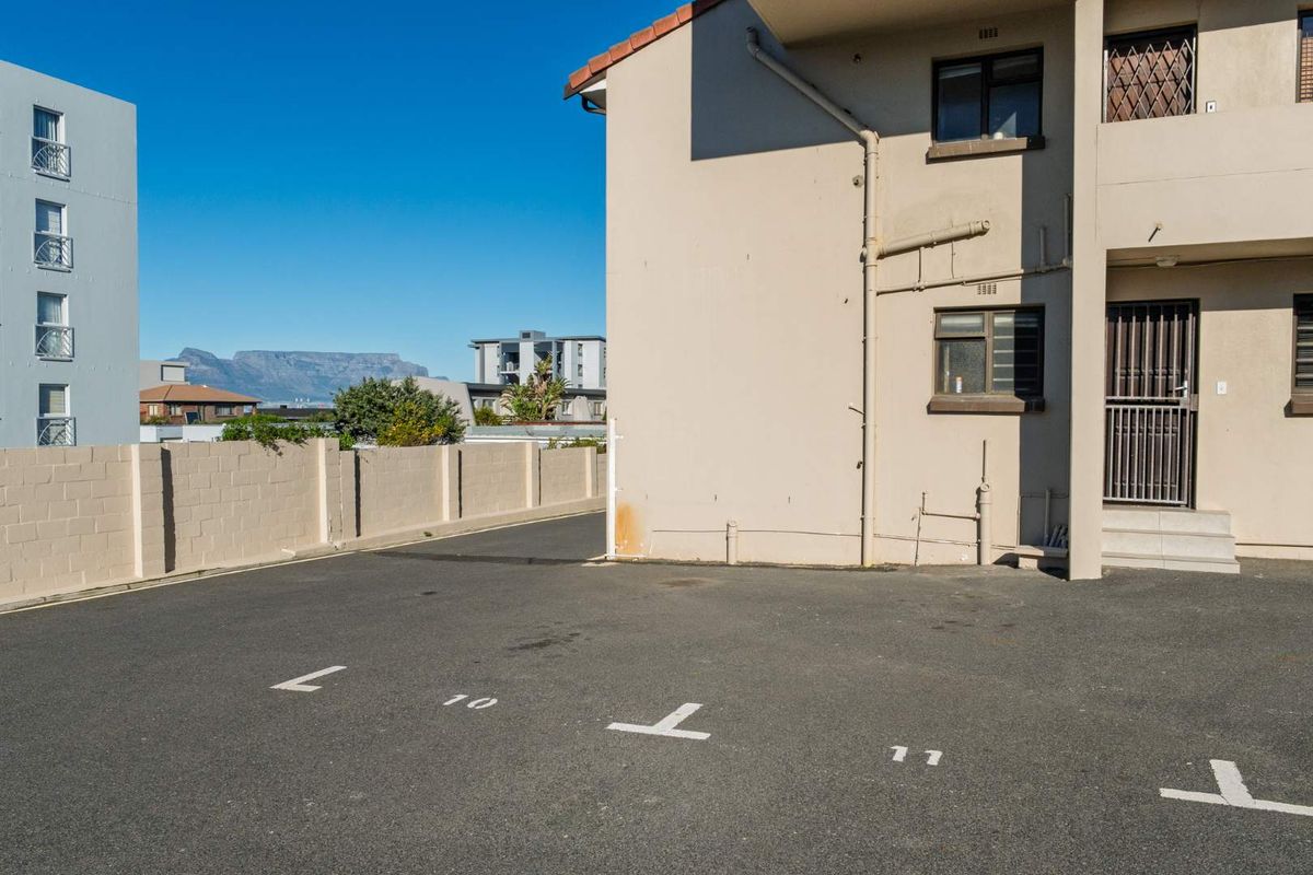 Exterior view of the apartment building showing the paved parking area, security gates, and surrounding neighbourhood with mountain views.