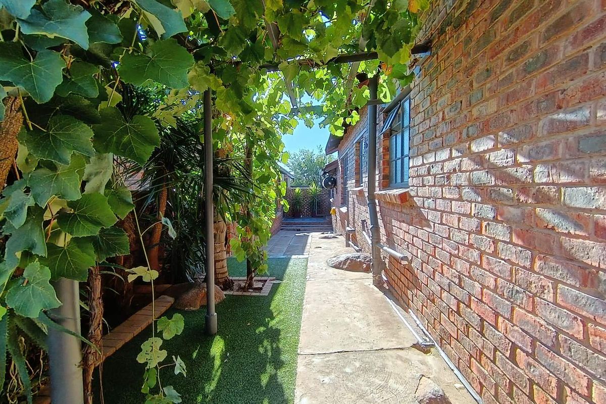 A side exterior walkway featuring a brick facade, paved path, artificial grass, and a vine-covered pergola structure.