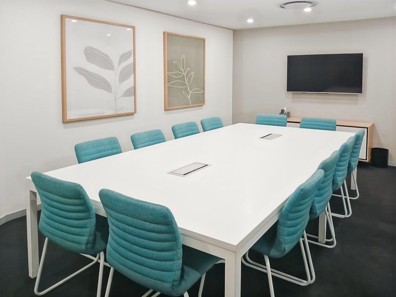 A modern, well-lit office boardroom featuring a large central meeting table, recessed ceiling lighting, and a wall-mounted television for presentations.
