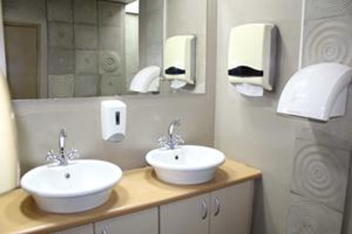 A clean and functional office bathroom featuring a dual vanity with white basins, wall-mounted dispensers, and a large mirror.