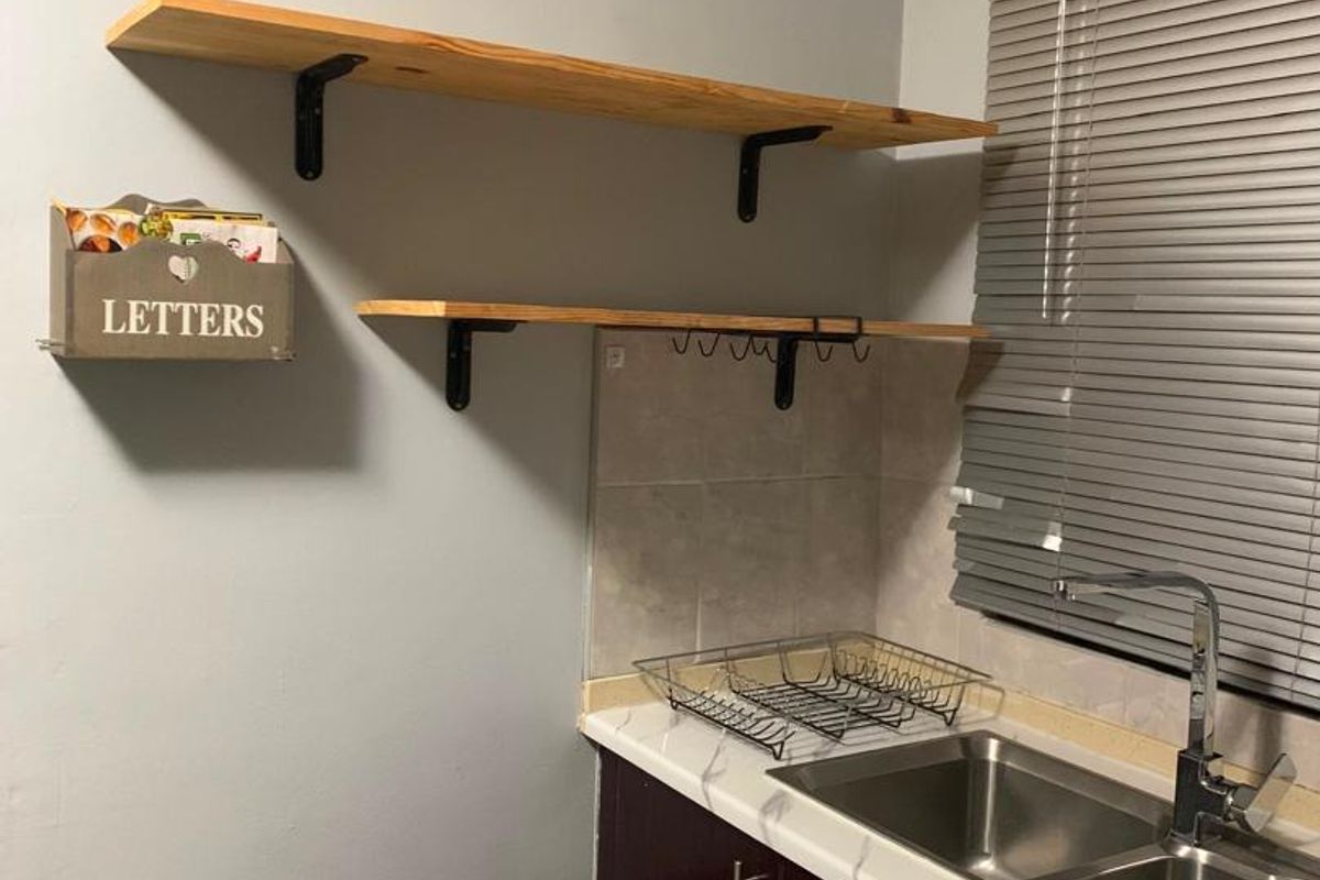 A compact kitchen area featuring dark cabinetry, a double stainless steel sink, marble-effect countertop, and open wooden shelving.