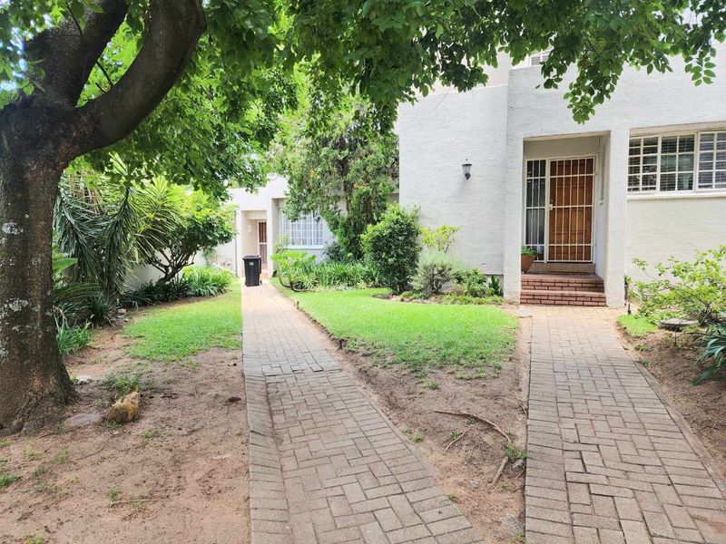 Front exterior view of the townhouse featuring a paved walkway leading to the entrance, surrounded by established garden beds and mature trees.