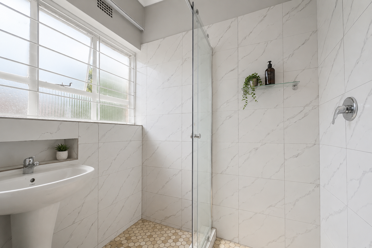 Modern bathroom featuring a pedestal sink, a glass-enclosed shower with marble-look wall tiles, and a window with security bars.