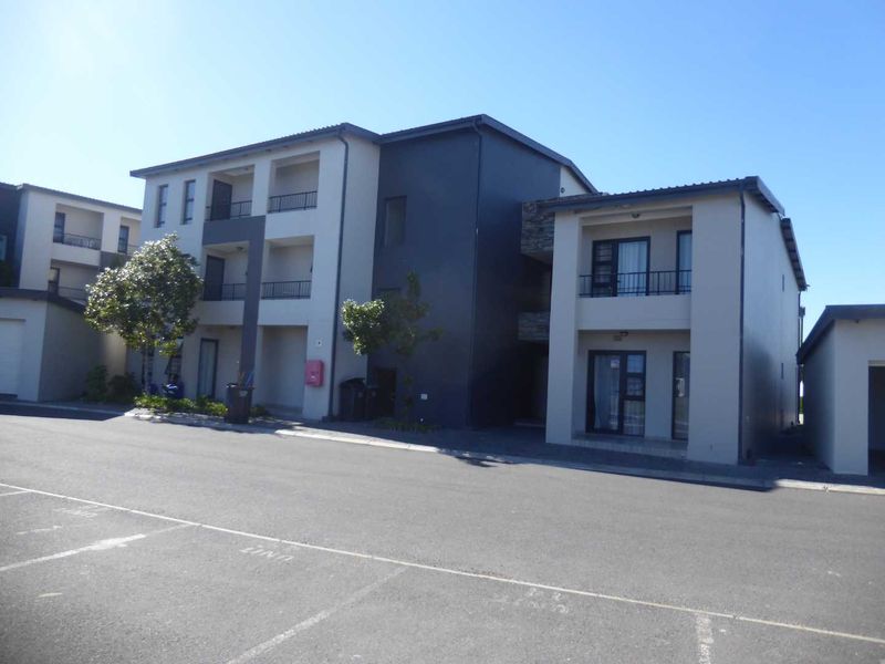 A modern multi-story apartment complex featuring a paved parking area, private balconies with metal railings, and a contemporary two-tone exterior finish.