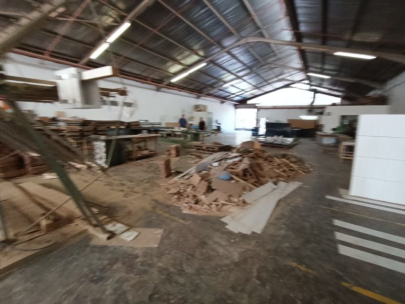 Large industrial warehouse space featuring high exposed steel truss ceilings, fluorescent lighting, and concrete flooring, currently utilized for storage and workshop activities.