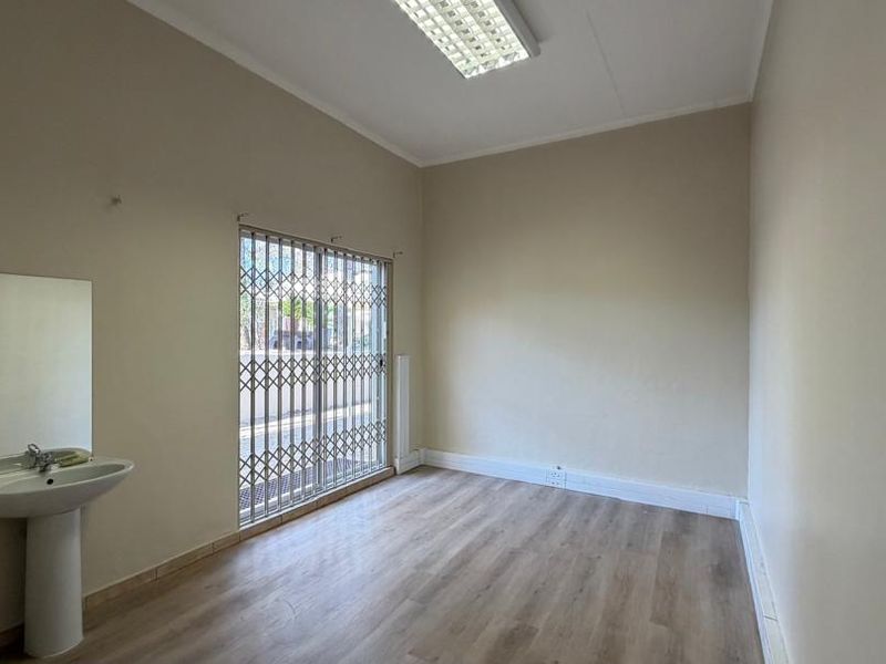 A clean, empty office space featuring laminate flooring, a built-in pedestal sink, and a secure sliding door with a metal security gate.