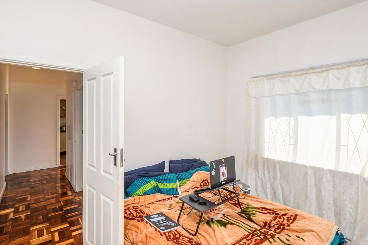 A bedroom featuring classic parquet flooring, a white panel door, and a window with security bars, offering a functional and well-lit living space.