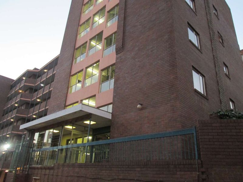 Exterior view of a multi-story apartment building featuring a brick facade, concrete entrance canopy, and secure metal perimeter fencing.