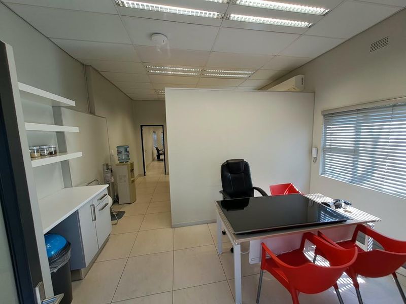 A functional office space featuring tiled flooring, a suspended ceiling with recessed lighting, a small kitchenette area, and an integrated air conditioning unit.