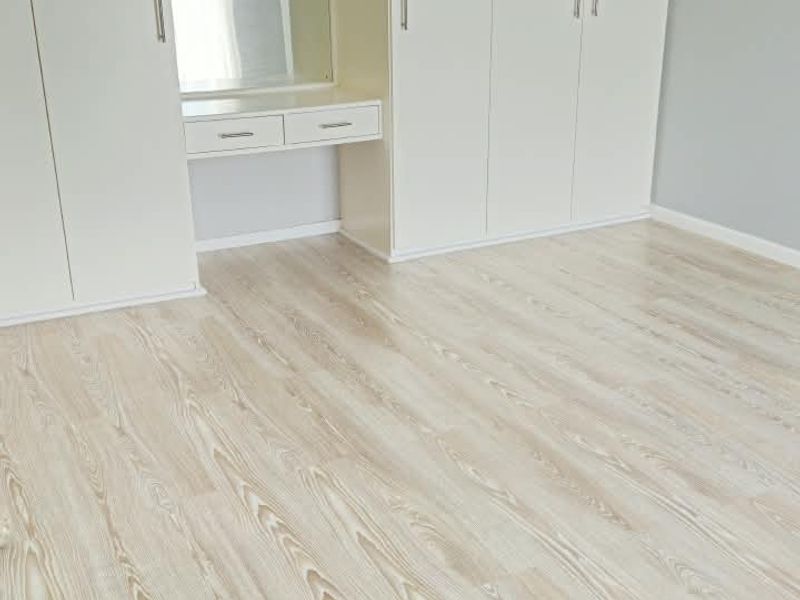 A bright, empty bedroom featuring light-toned laminate flooring, extensive built-in cabinetry with an integrated vanity, and exposed white ceiling beams.