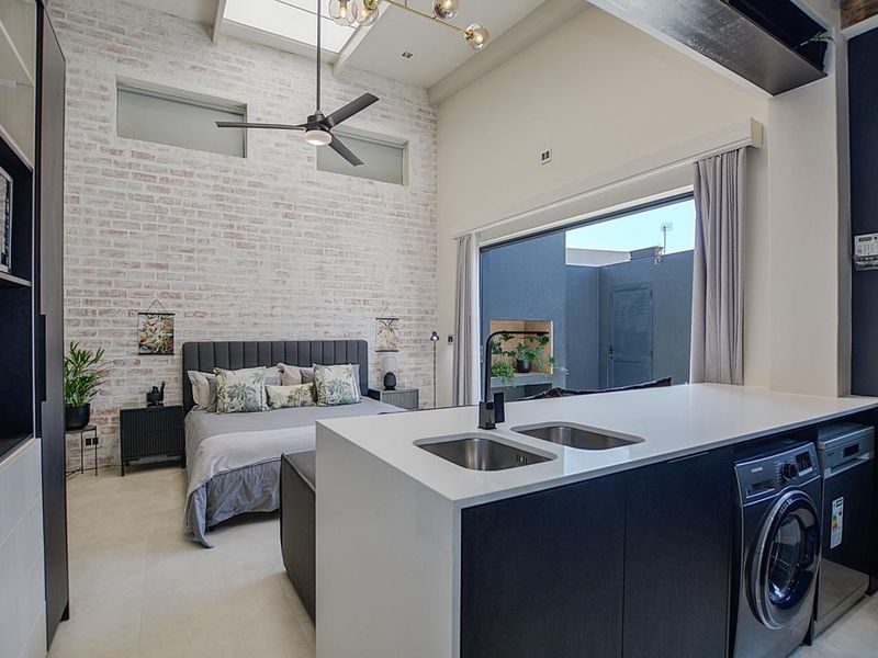 Modern open-plan studio apartment featuring a high-ceiling exposed brick wall, skylight, and a sleek kitchen island with integrated appliances and direct patio access.