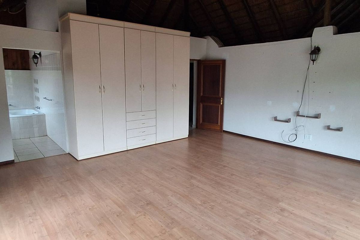 Spacious bedroom featuring laminate flooring, built-in cupboards, and an en-suite bathroom, set under a high vaulted thatched ceiling.