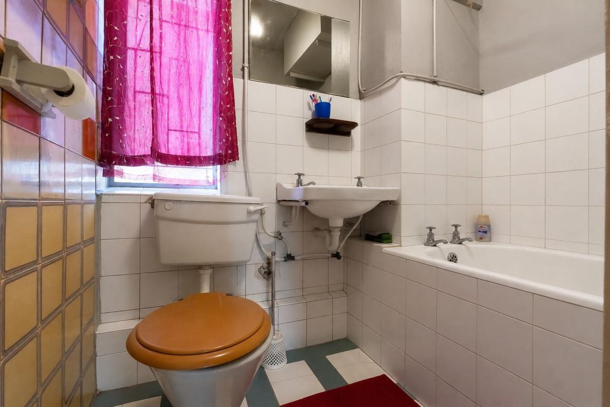 A compact, dated bathroom featuring a standard bathtub, wall-mounted basin, and toilet, with original tiling and exposed plumbing fixtures.